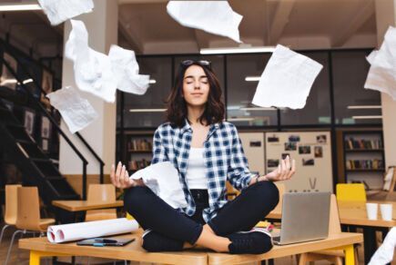 Jeune femme assise en tailleur sur son bureau avec ses dossiers qui volent pour symboliser la réduction de sa charge mentale.